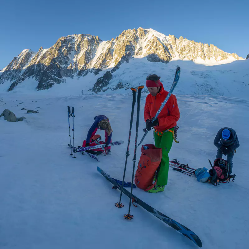 Drie mensen maken bij zonsopgang hun skiuitrusting klaar op een besneeuwde berghelling, met op de achtergrond hoge, zonovergoten bergtoppen en rugzakken en skistokken op de grond.